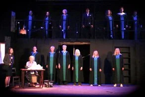 Scene from a stage play with six people in working robes in the foreground and five people dressed in formal robes standing on an elevated platform in the background. The scene involves a theatrical or academic presentation, with props including a de