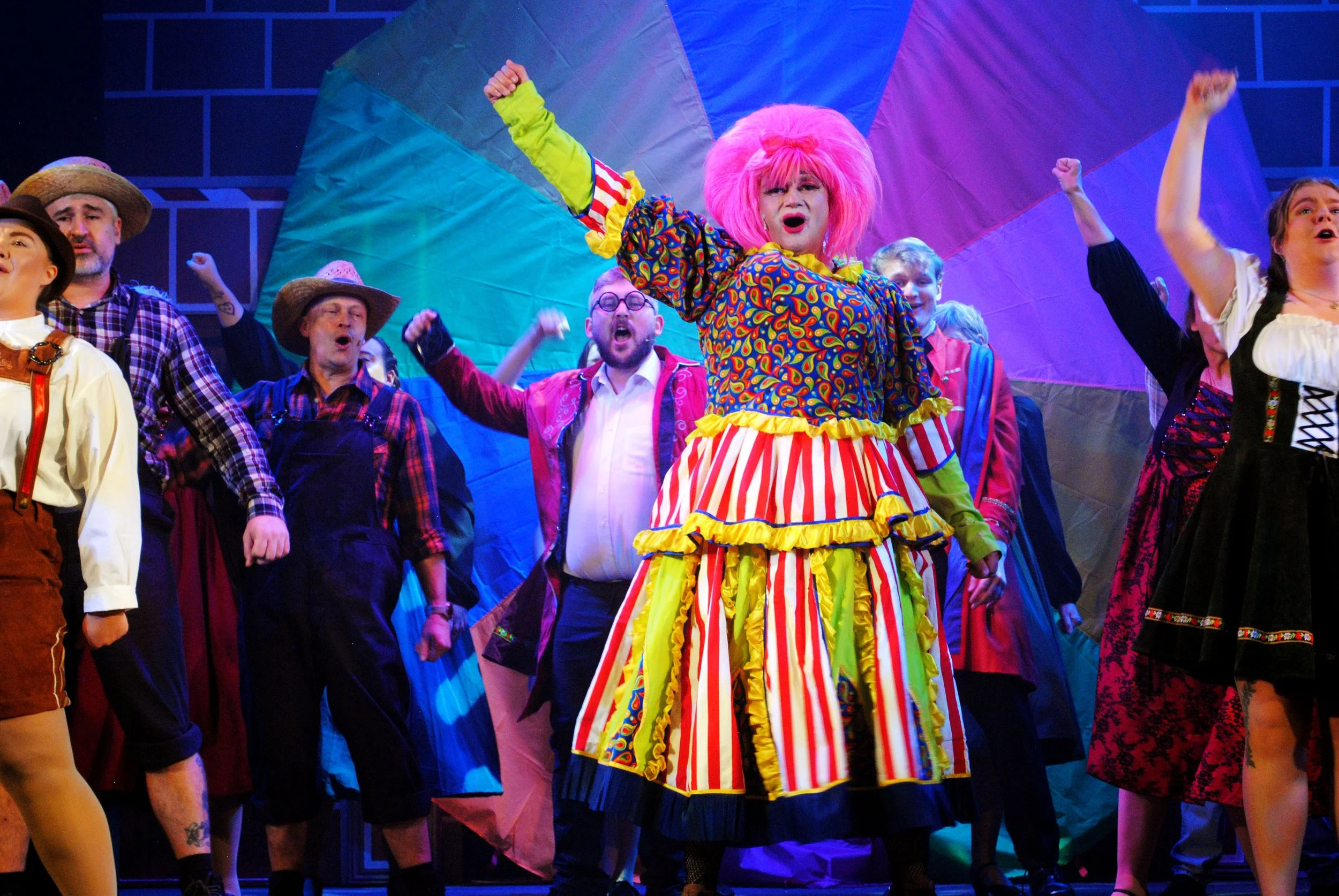 A colorful stage scene from a theatrical production featuring a woman dressed as a clown with pink hair, a multicolored dress with red, white, and yellow stripes, surrounded by actors in period costumes, with a vibrant background.