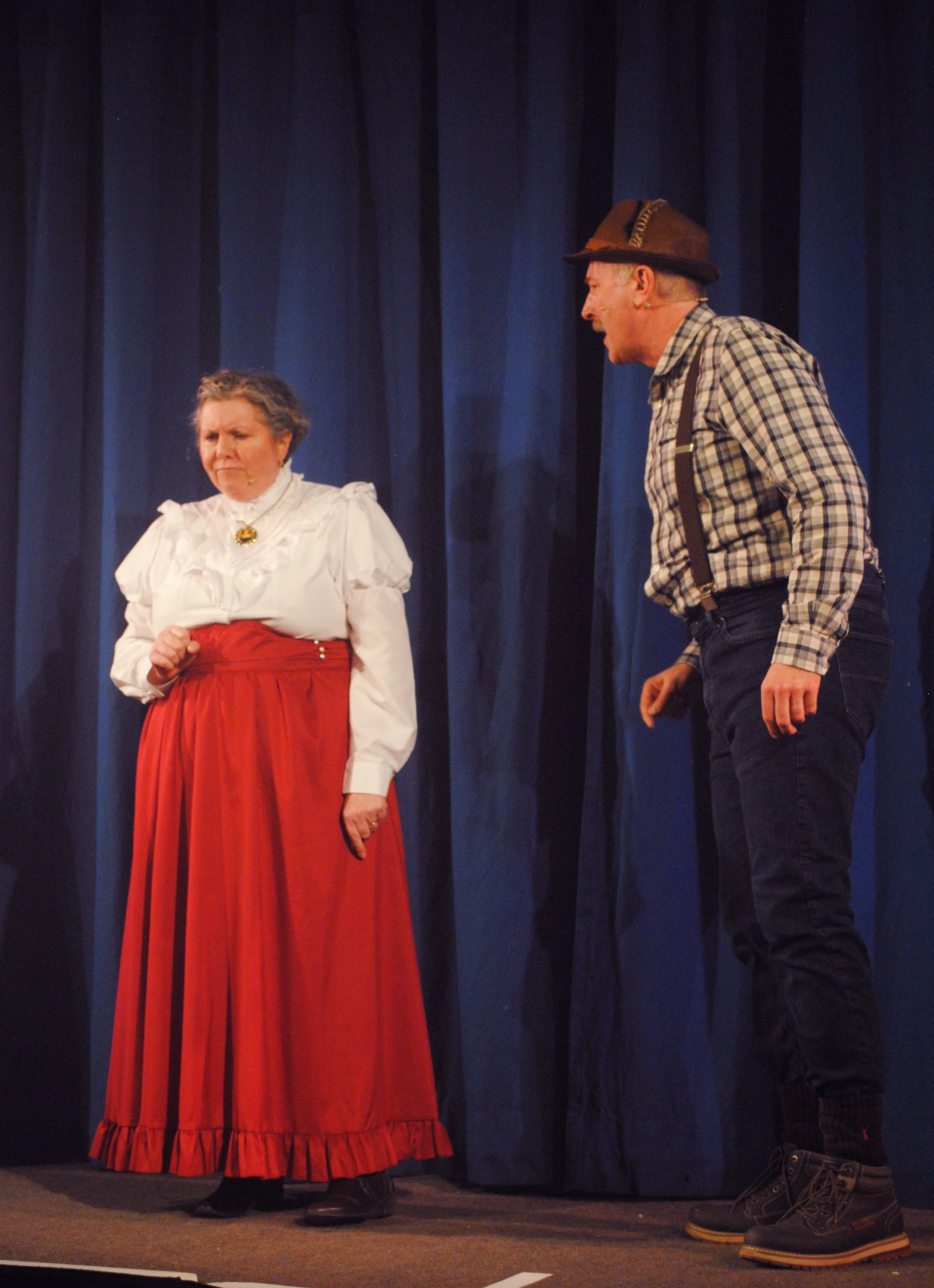 Two actors on stage, one woman and one man, performing in a play. The woman is wearing a white blouse with ruffles and a long red skirt, looking displeased. The man, wearing a plaid shirt, suspenders, and a hat, is leaning slightly towards her, speak