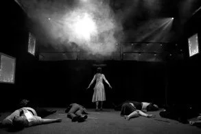 A woman stands on stage with her arms outstretched, while four people lie on the floor in front of her in a dark theater or performance space.