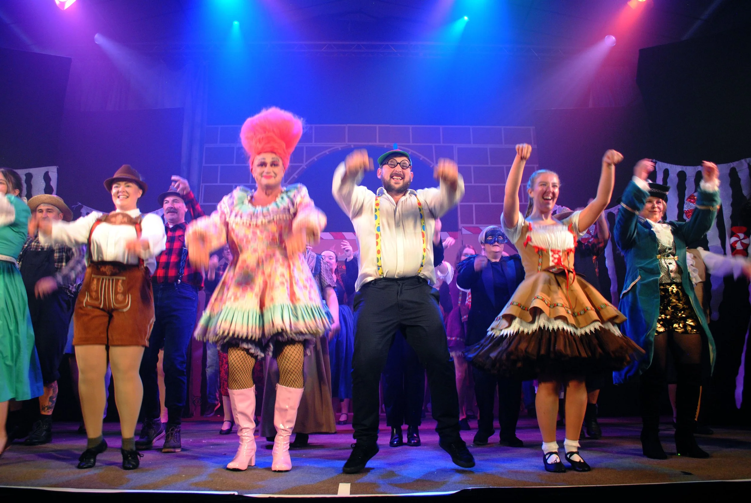 The image depicts a lively stage performance with a diverse cast of actors dressed in colorful costumes, dancing and celebrating under vibrant stage lighting.