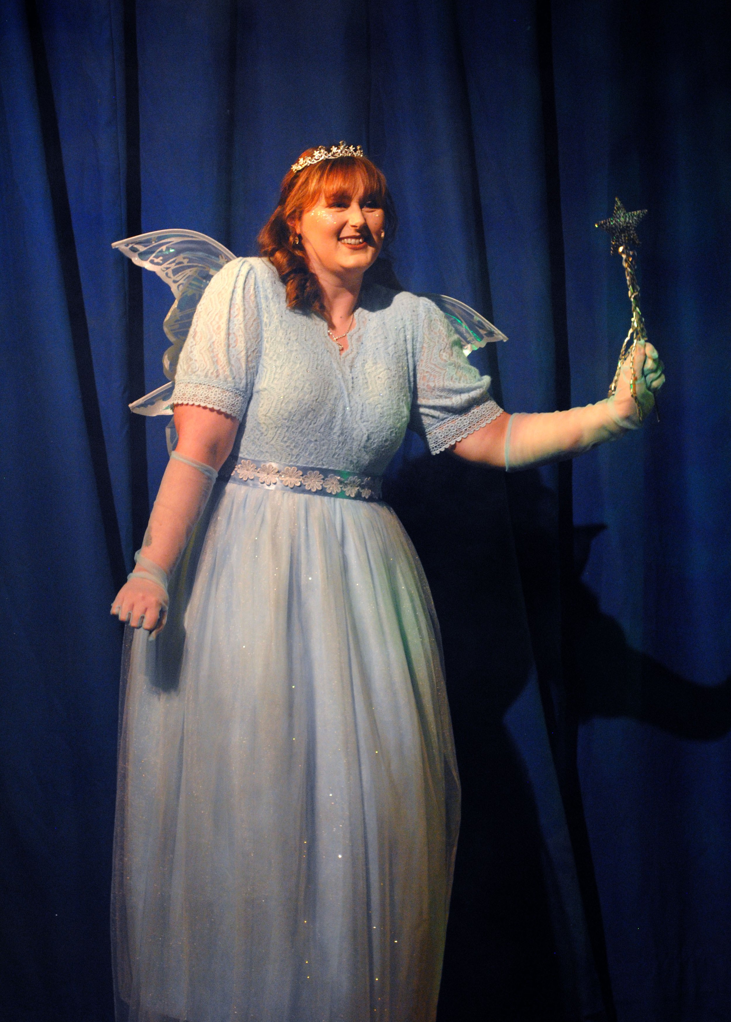 Woman dressed as a fairy or princess, wearing a light blue gown, fairy wings, a tiara, and holding a star wand, standing in front of blue curtains.