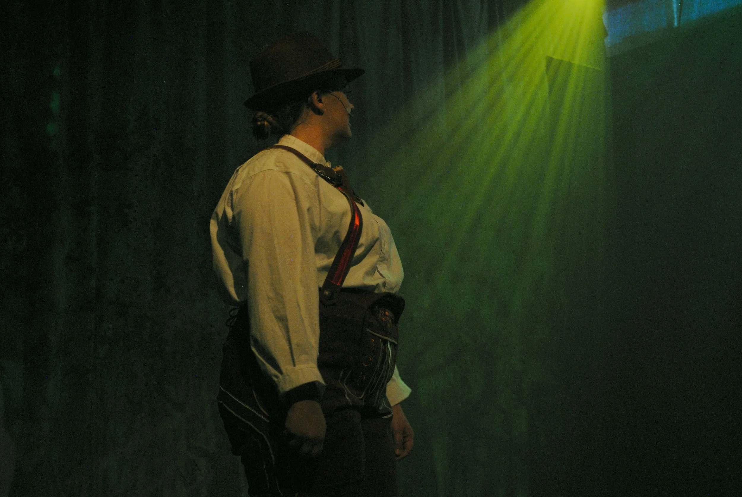 Person dressed in traditional German attire, including a hat, white shirt, suspenders, and dark pants, standing under green stage lighting in a dimly lit setting.
