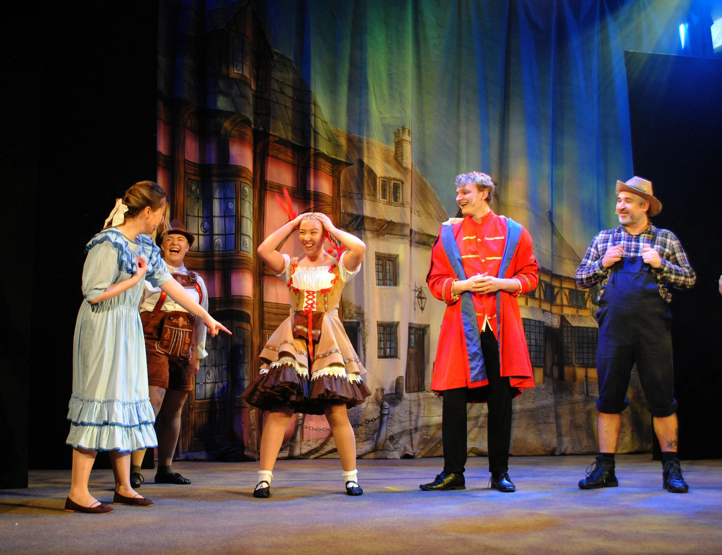 Actors performing on stage in colorful costumes, with a backdrop of a town scene. One actor in a blue dress points and laughs, others smiling and engaging in the act.