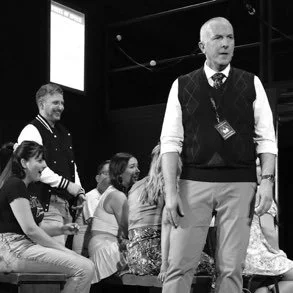 A man stands in front of a group of children and another man, in a room with a large screen in the background.