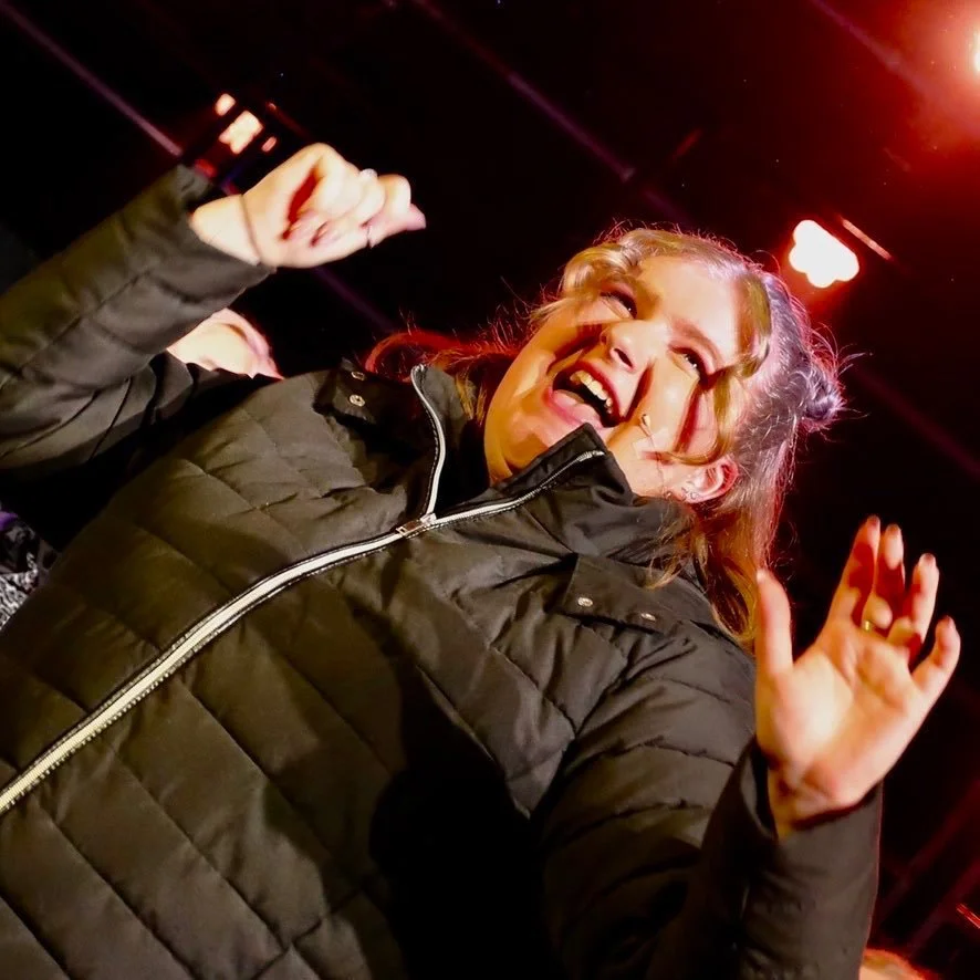 A young girl smiling and waving, wearing a black puffer jacket with a zip, in a dimly lit indoor space with red lighting.
