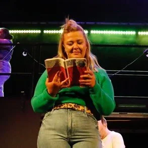 A woman with blonde hair, wearing a green sweater and jeans, reading a book.
