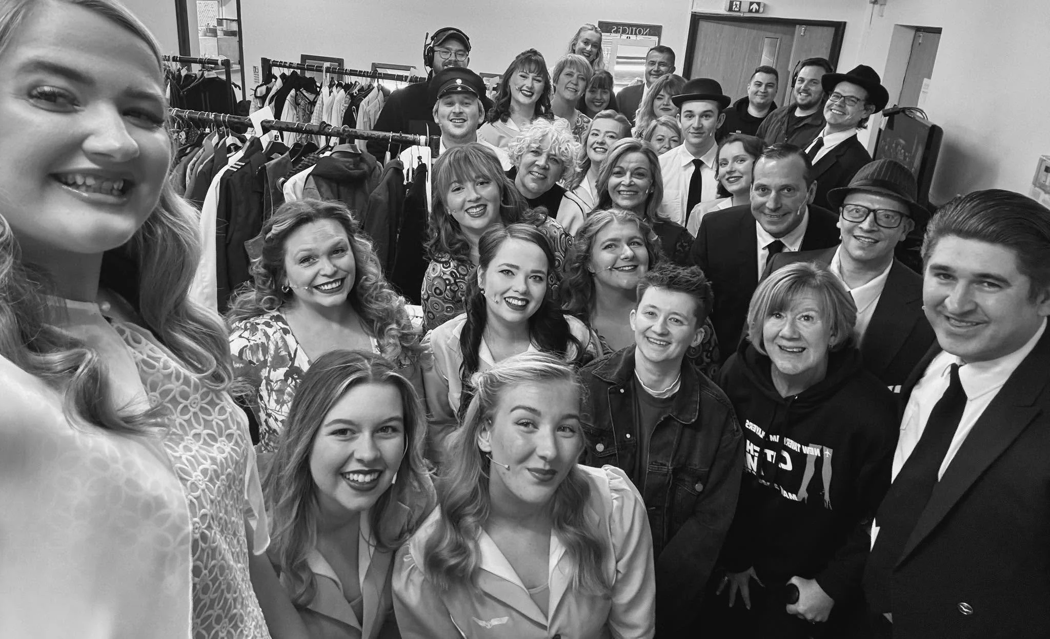 Black and white photo of a group of people posing together indoors, with clothing racks in the background, some wearing vintage or theatrical costumes.