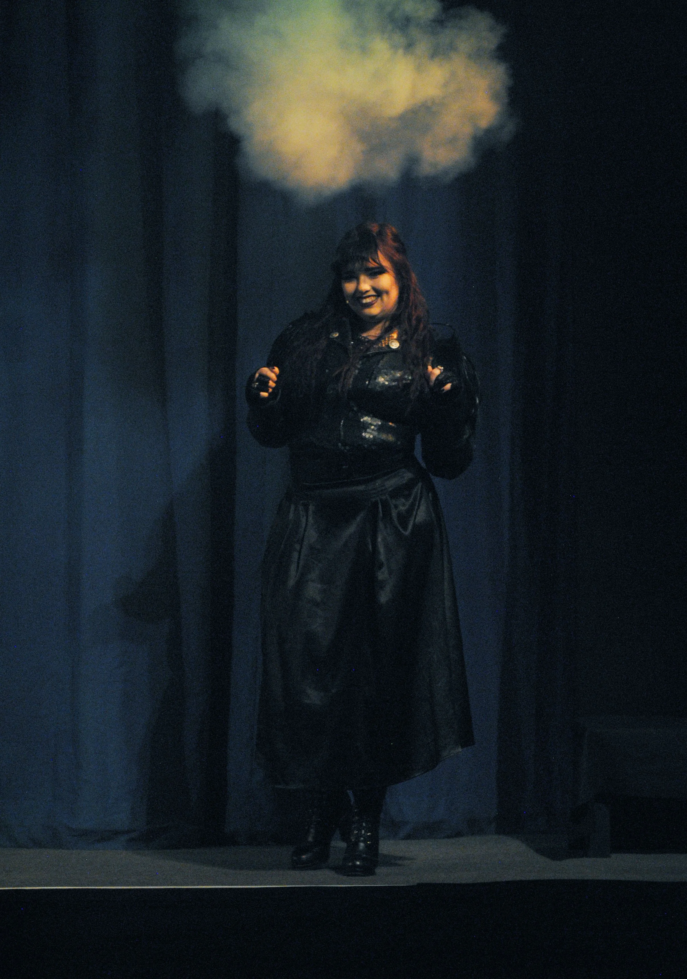 A woman stands on stage with a cloud of smoke above her head. She is wearing a black outfit and is smiling.