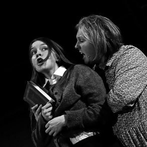 Two women engaged in a theatrical performance, with one holding a book and the other reacting expressively, in black and white.