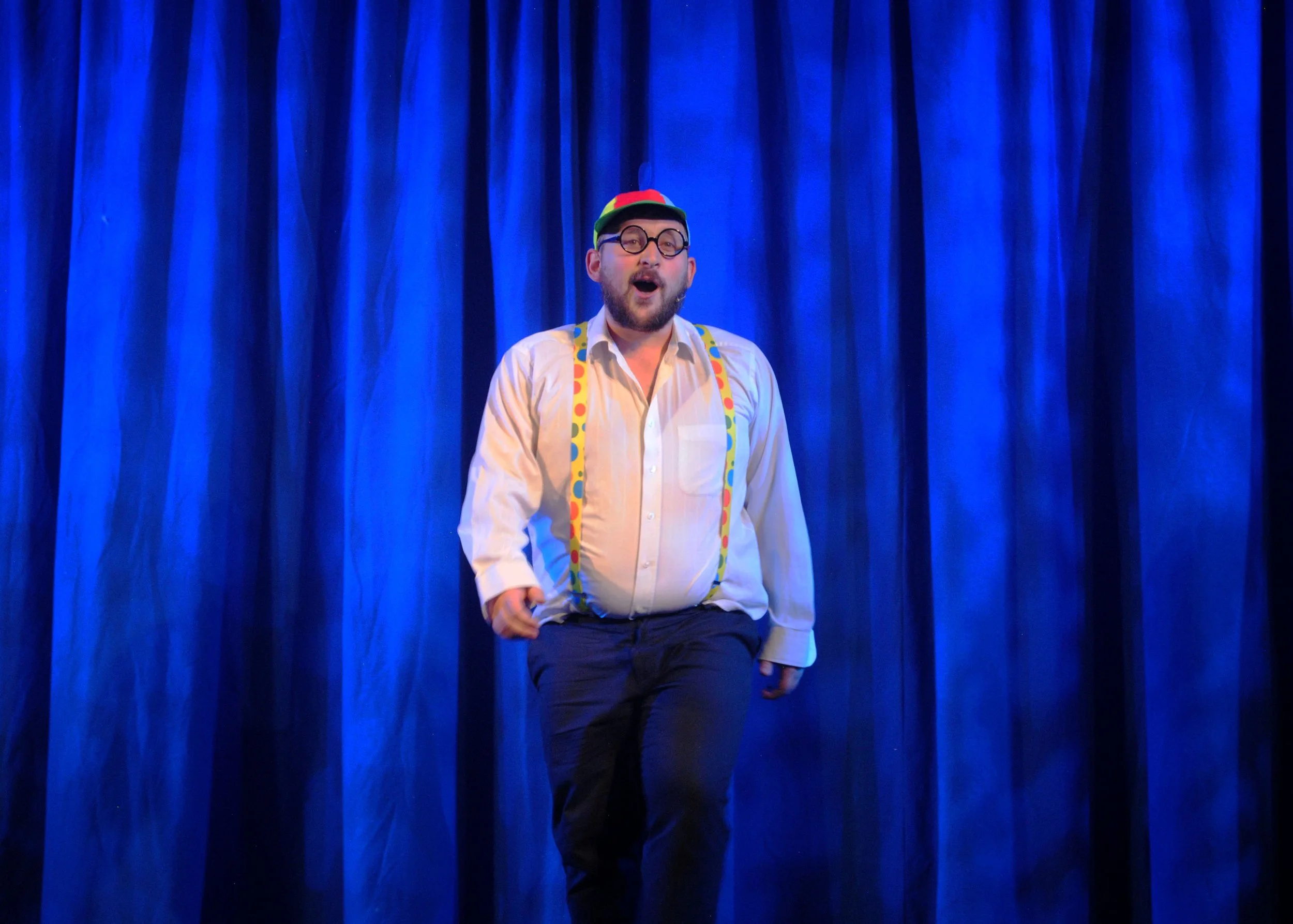 A man performing on stage in front of a blue curtain, wearing glasses, a hat, suspenders, and a white shirt.