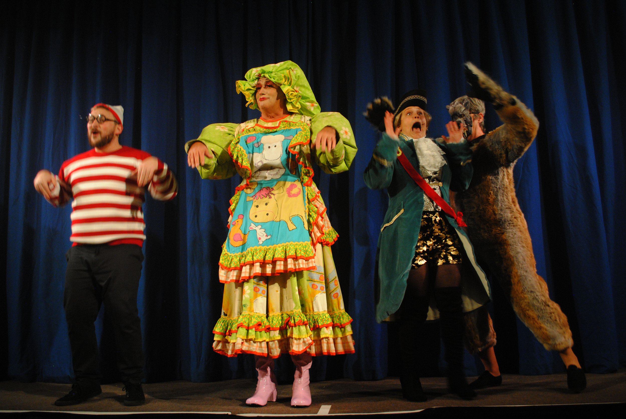 Four performers in colorful costumes on stage, with blue curtain backdrop, engaging in a theatrical performance.