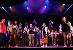 A stage with a group of people performing a musical or play, with musicians in the background and actors in costumes, under stage lighting.