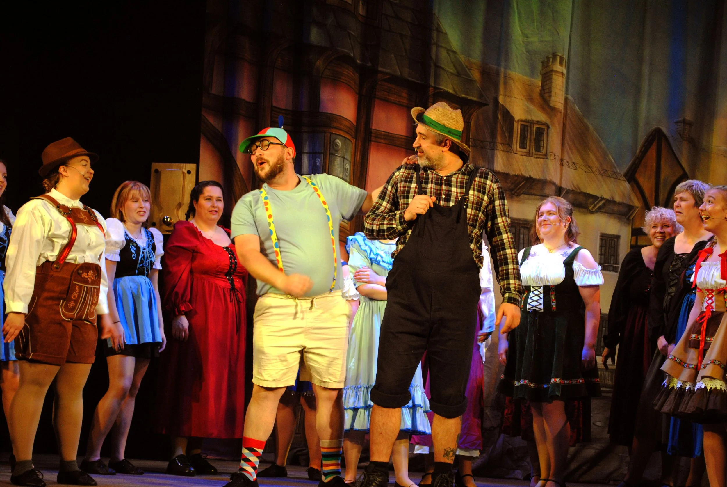 A stage scene from a theatrical performance featuring actors in costume, with colorful costumes and set design depicting a village or town.