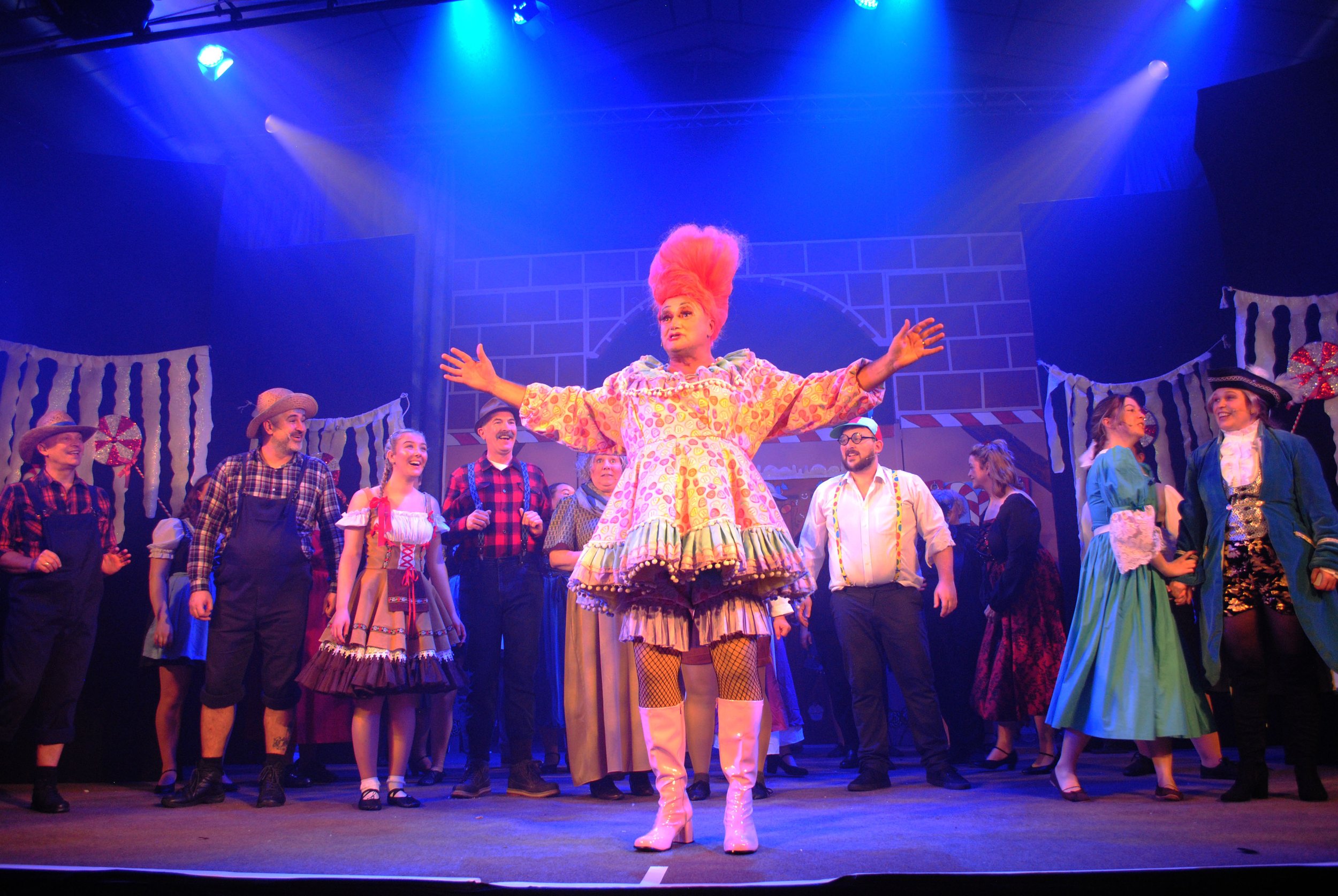 A theatrical stage performance featuring a woman with bright pink hair in a colorful dress, standing with arms outstretched. She is surrounded by cast members in period costumes, including cowboy hats, plaid shirts, and dresses, under purple and blue