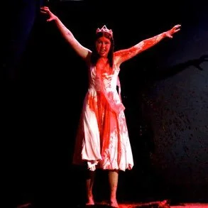 A woman wearing a crown and a blood-stained dress with arms outstretched on stage.