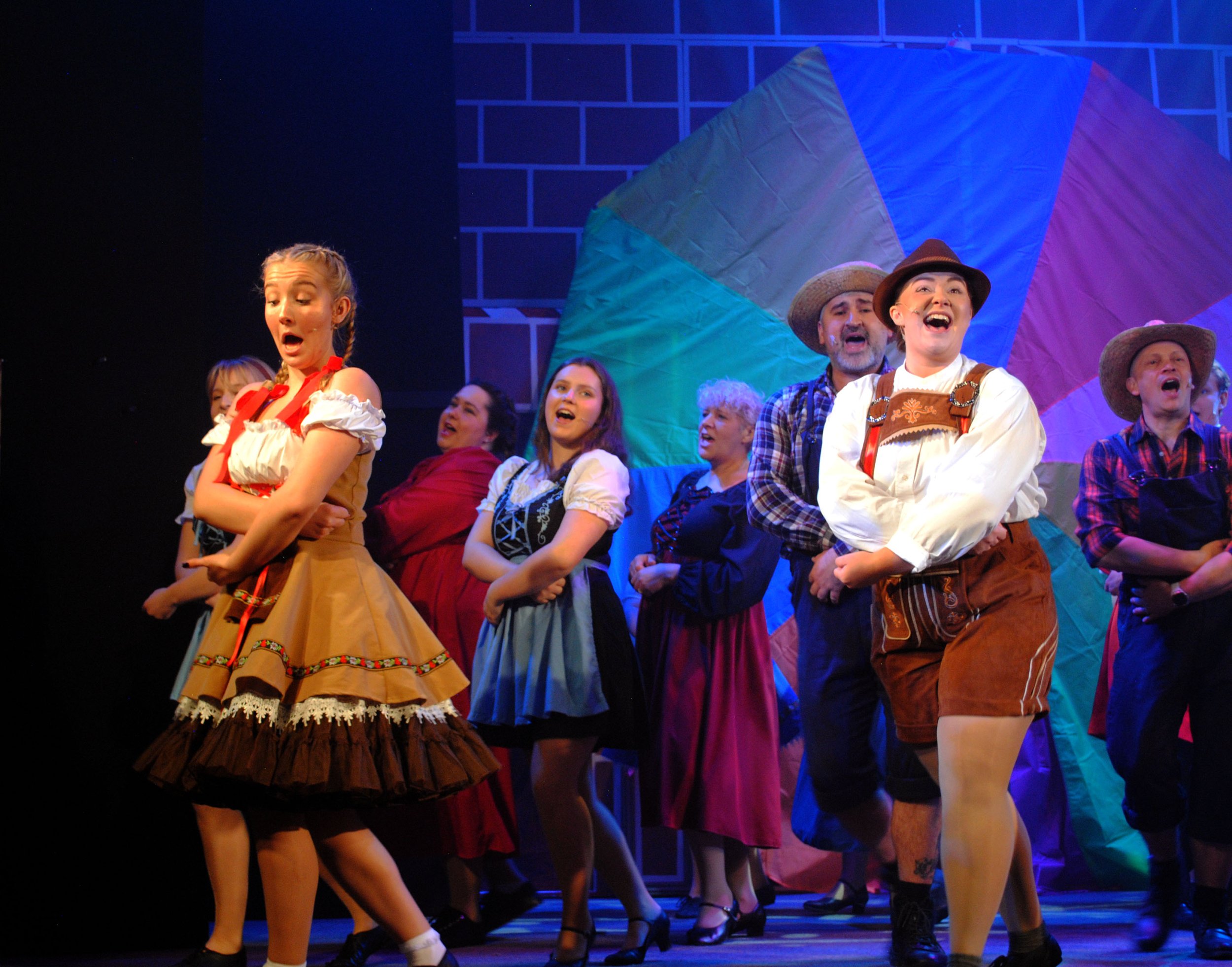 Group of performers on stage dressed in colorful, traditional, folk costumes, appearing to sing or act during a theatrical performance, with a large rainbow-colored umbrella backdrop.