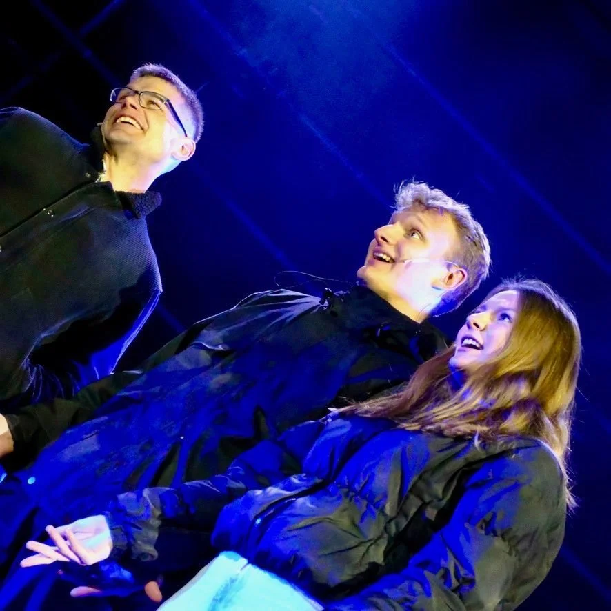 Three young adults standing on a stage, smiling and looking at each other with a blue stage light shining overhead.