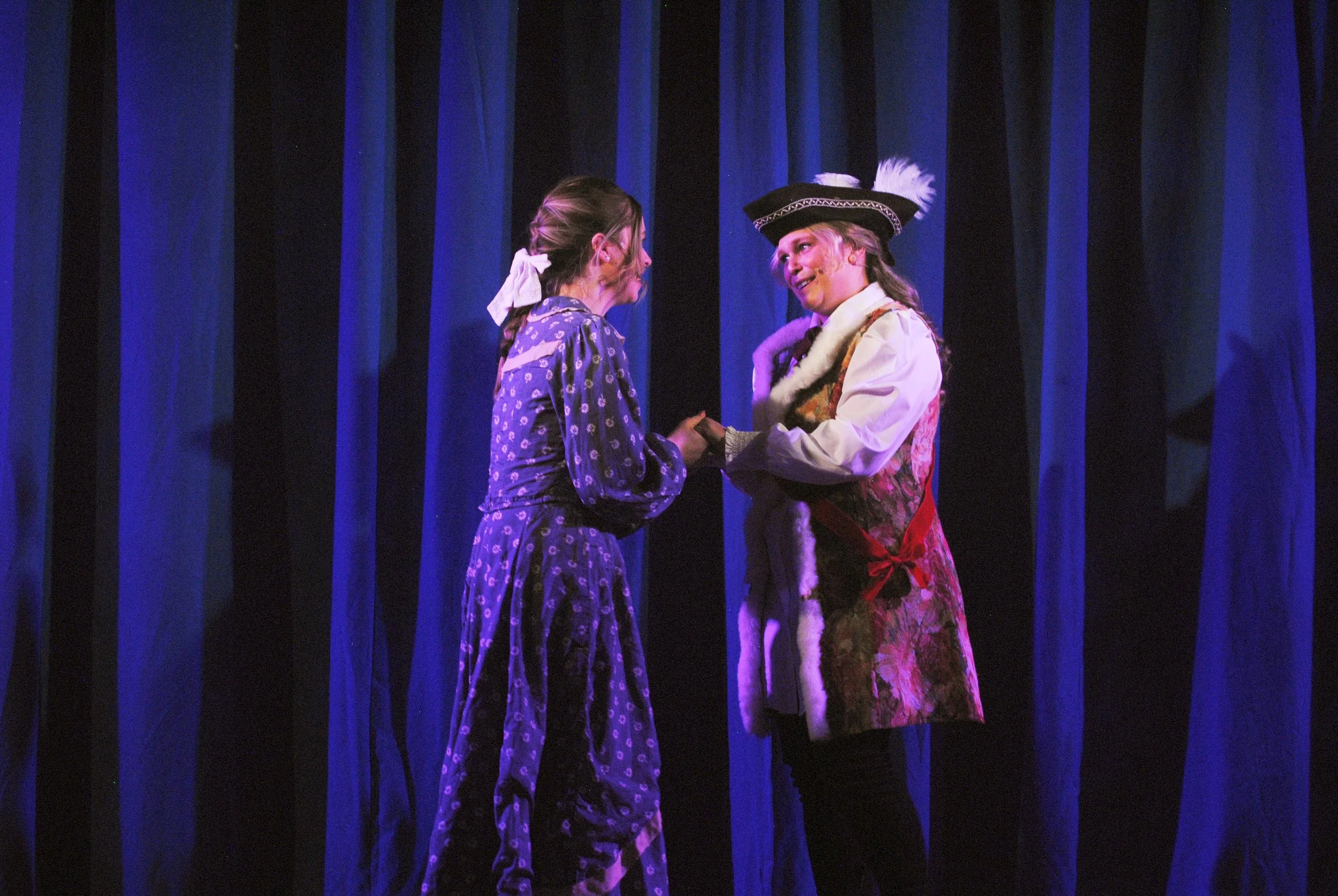 Two women dressed in period costumes stand on a stage with blue curtains, holding hands and talking.