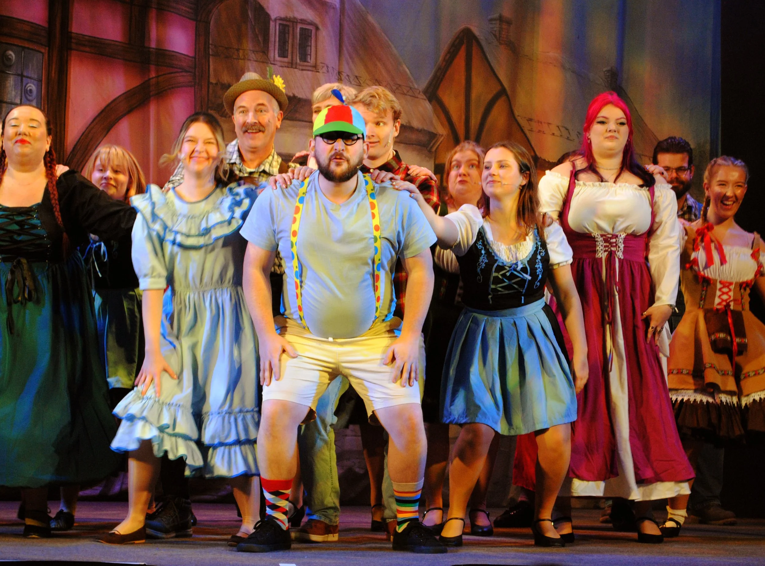 A group of actors on stage during a theatrical performance, dressed in colorful costumes, with some smiling and interacting, and a painted backdrop of a town scene in the background.