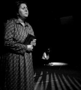 A woman standing on stage holding a book or notebook, with a spotlight on her and a dark background.