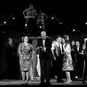 Group of people on stage during an award ceremony or event, with some individuals holding awards and others smiling for the camera, and a couple of performers or speakers in the background.