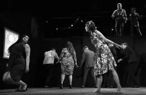 People standing in a circle with a woman dancing in the center at an indoor gathering.