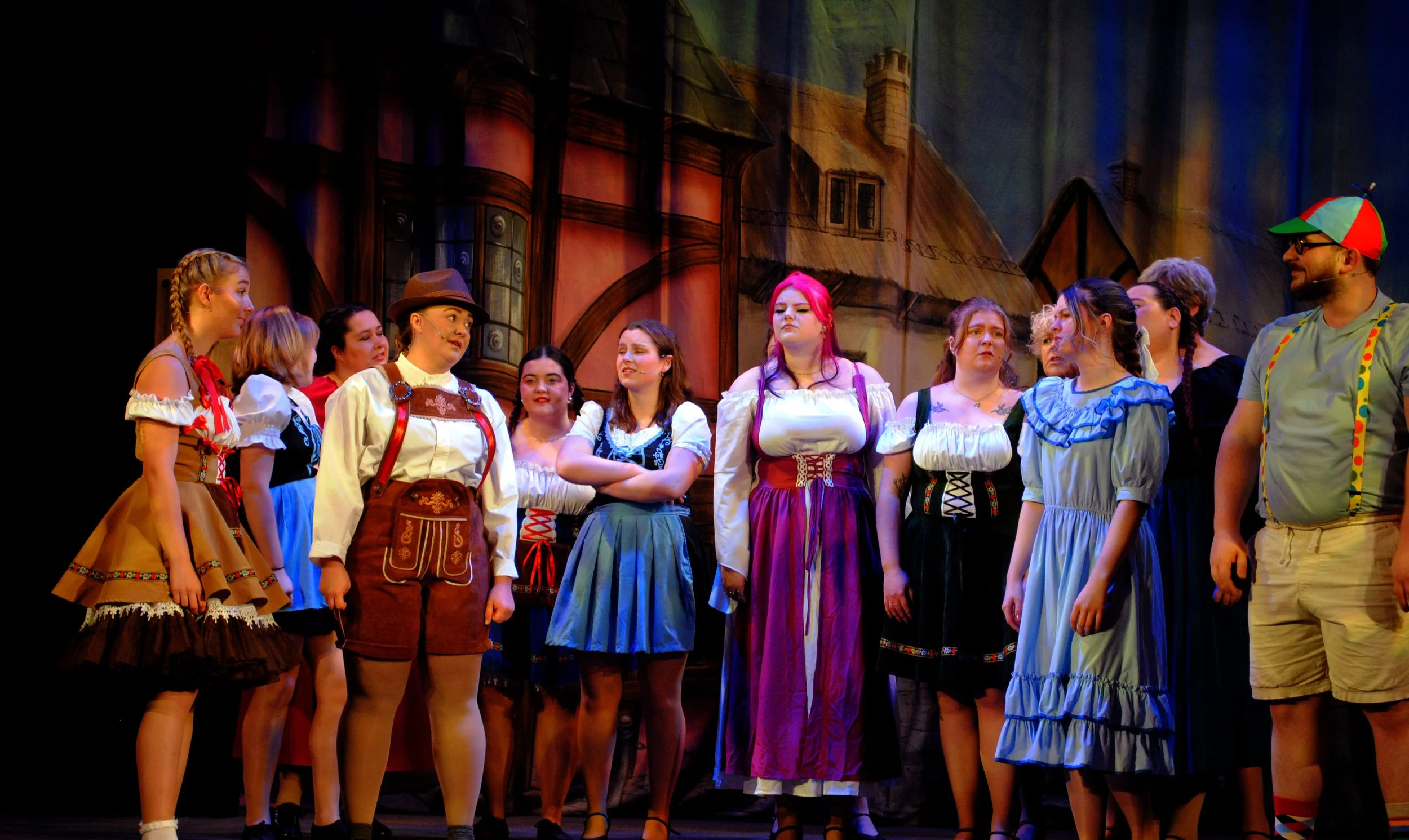 A group of people in colorful costumes on stage during a theatrical performance, with a painted backdrop of a house and street scene.