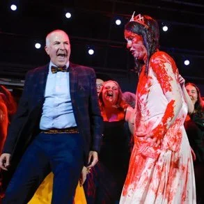 A man and a woman dressed as a bloody bride on stage with surprised or shocked expressions, surrounded by a few other women.