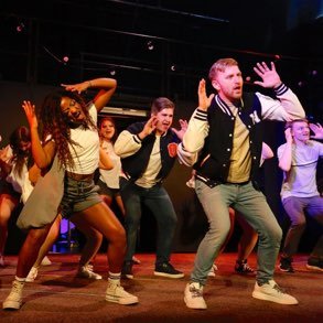 A group of people dancing on stage during a performance or rehearsal, with energetic movements and focused expressions.