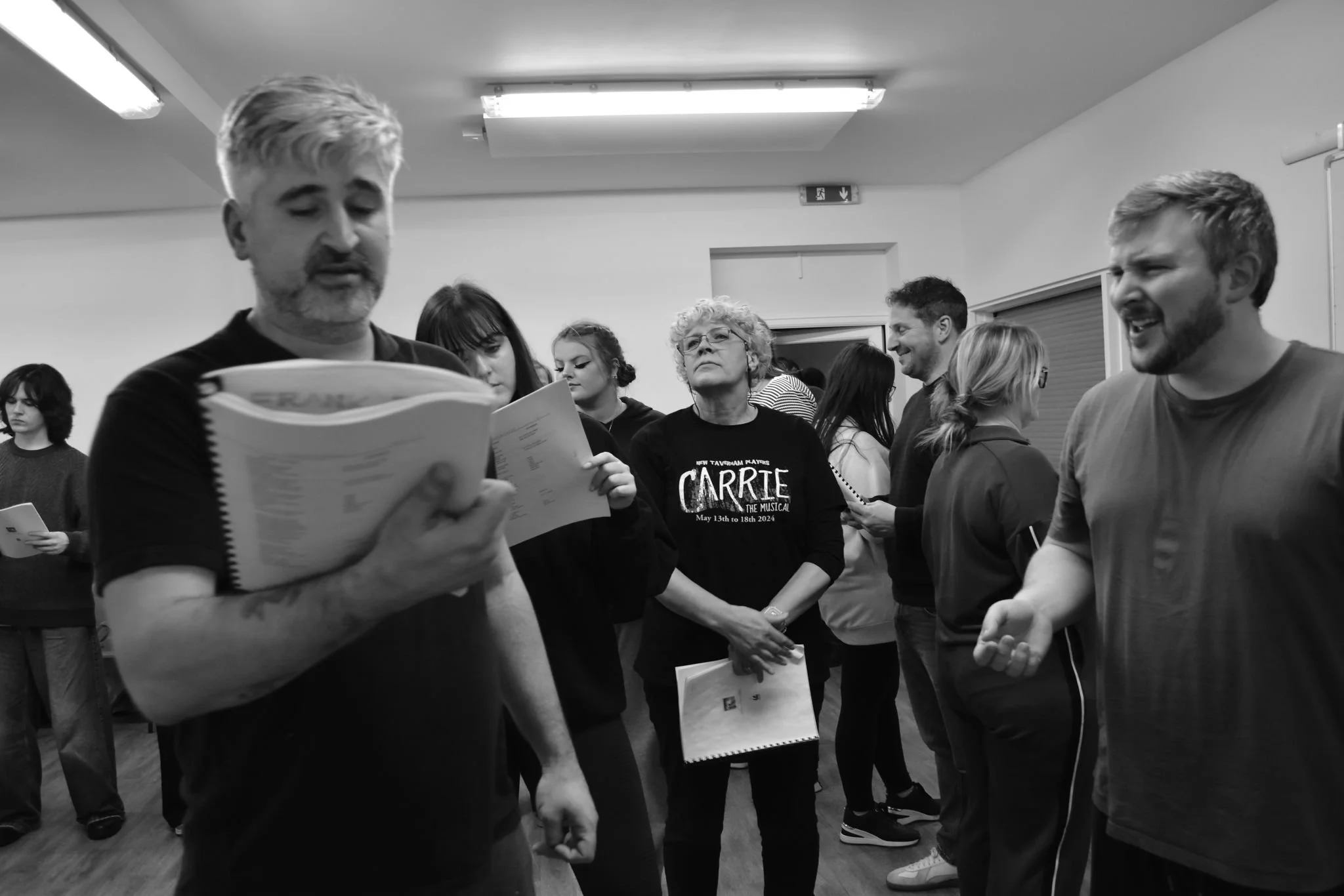 Group of people in a room, some reading from scripts or notes, engaged in a rehearsal or practice.