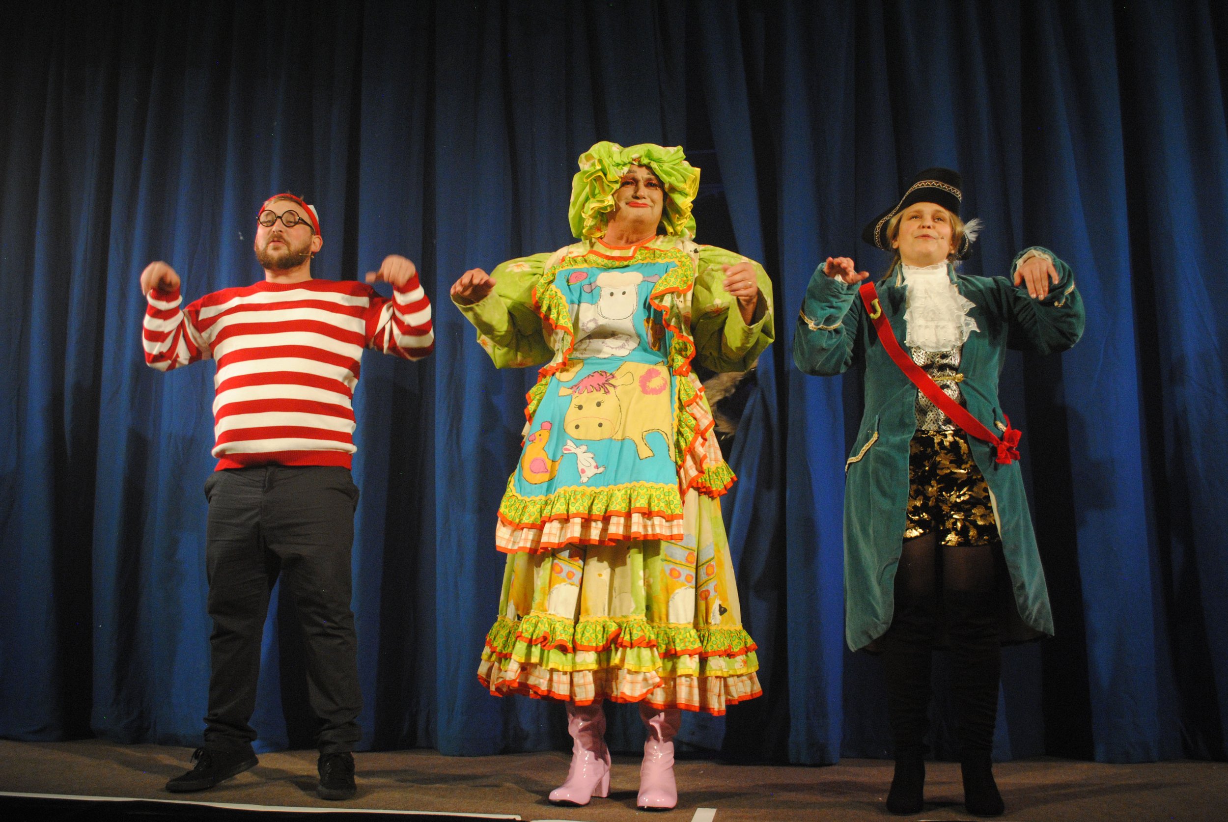 Three performers on stage dressed in colorful costumes, standing in front of a blue curtain. The person in the middle wears a vibrant, cartoon-themed dress with green hair, pink boots, and a cheerful expression. The person on the left wears a red and