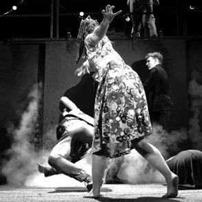 A woman in a floral dress with an umbrella over her head dancing with a man on a stage, with smoke and other people in the background.