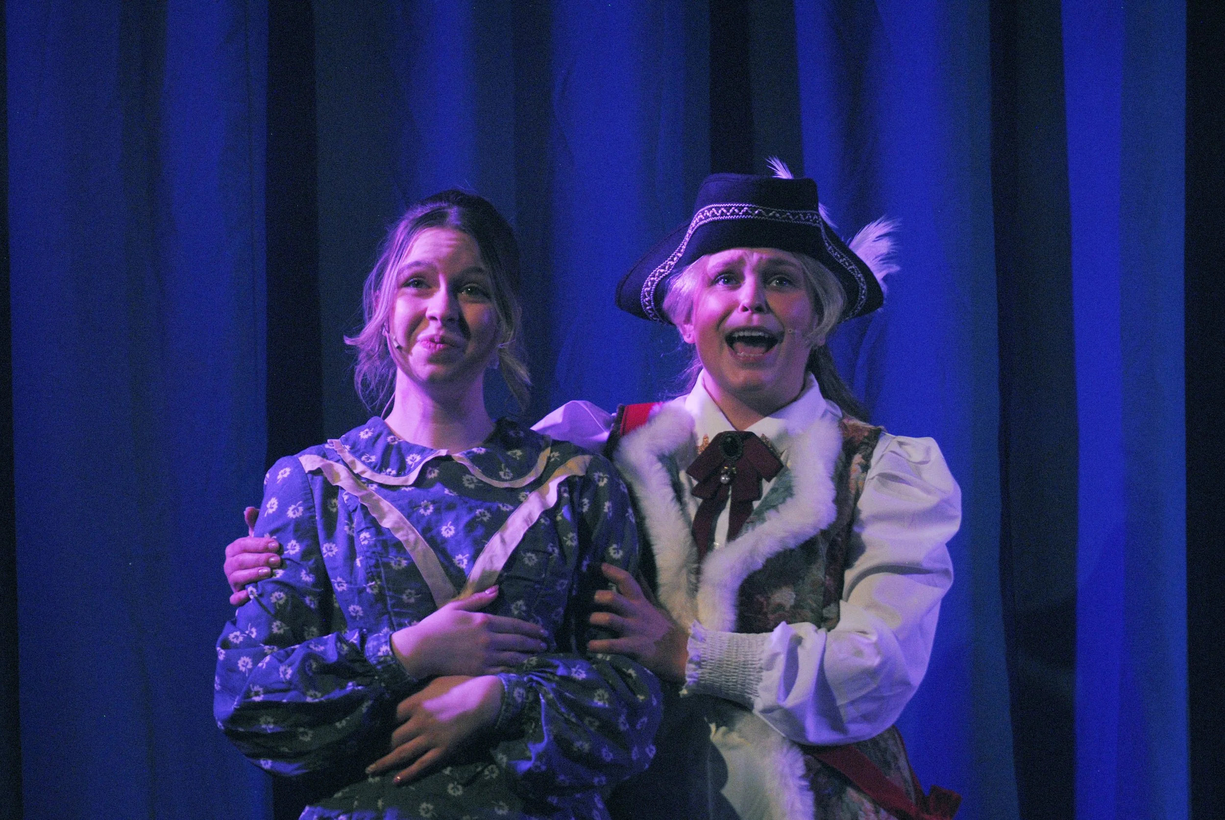 Two women dressed in theatrical costumes on stage, with blue stage curtains in the background. The woman on the right is wearing a historical costume with a hat and fur trim and appears to be singing or speaking passionately, while the woman on the l