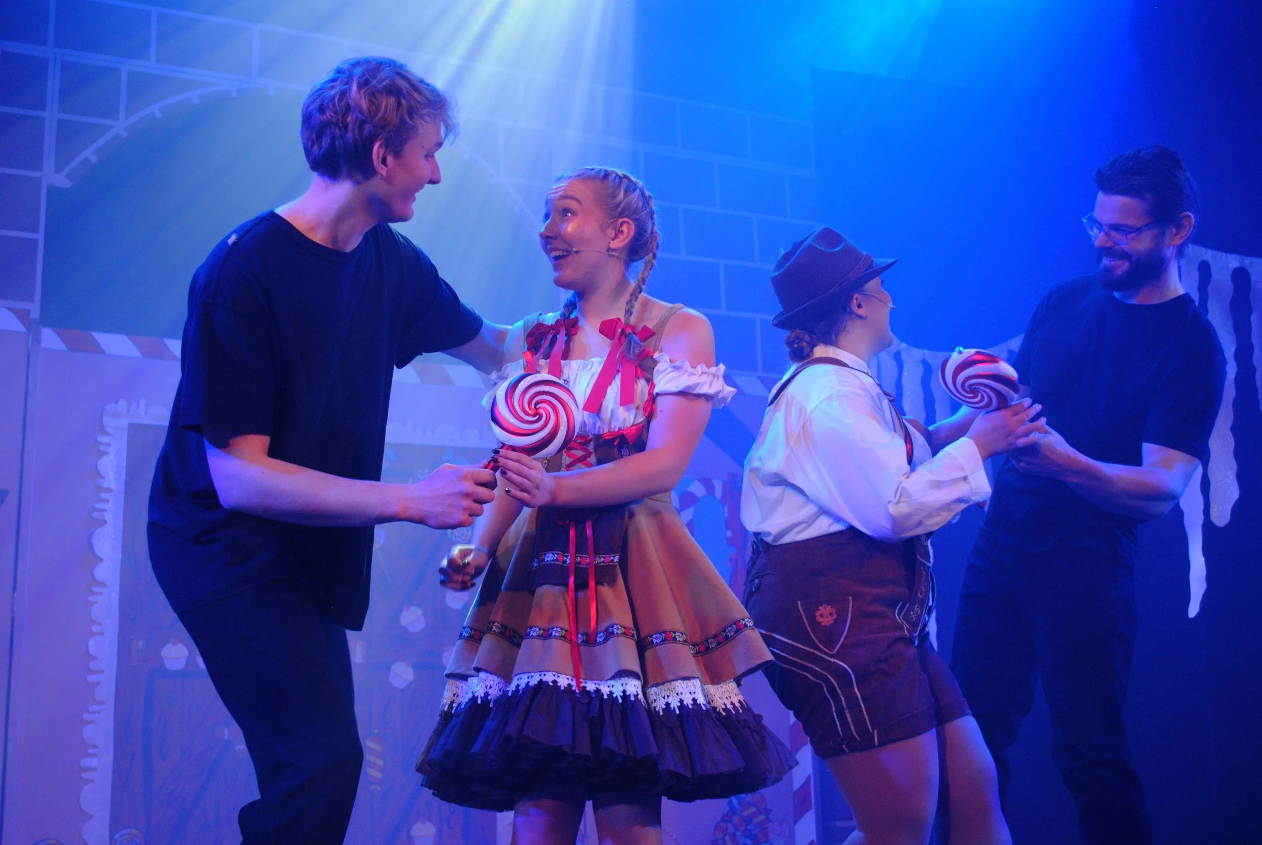 People on stage sharing large lollipops, engaging in conversation, with colorful stage background.