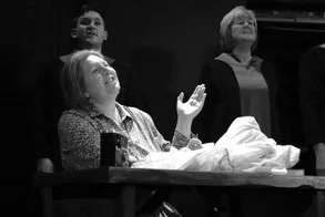 Three women in a room, one seated at a table with her hand raised, and two standing behind her, all appearing focused or engaged in conversation.