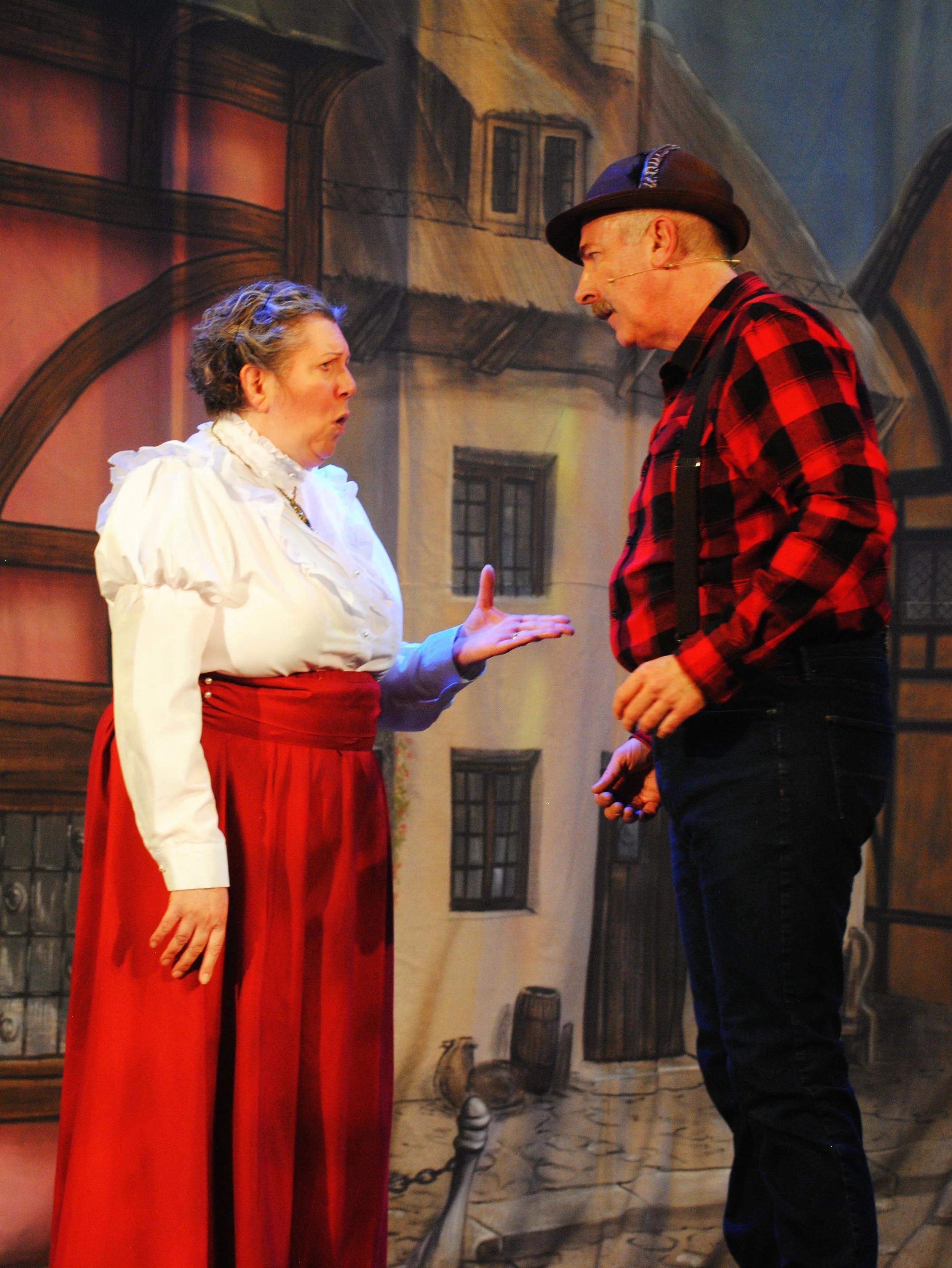 Two actors, a woman and a man, are performing on stage. The woman has curly hair, is wearing a white blouse with puffed sleeves, a red skirt, and has an expressive face. The man is wearing a black hat, a red and black checkered shirt with suspenders,