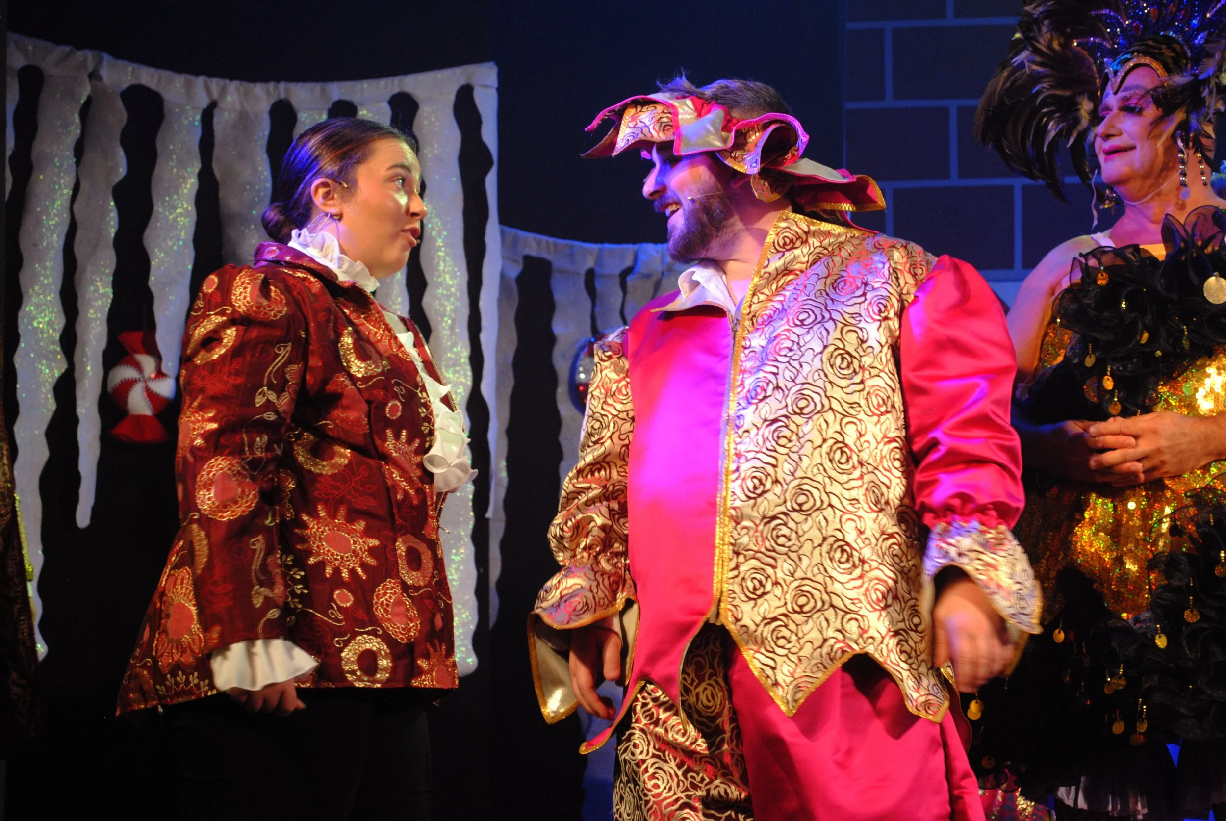 Three actors in colorful, elaborate costumes on stage, performing in a theatrical play with a black and white backdrop decorated with stockings.
