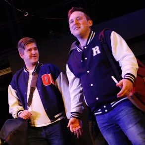 Two young men wearing letterman jackets standing on a stage or in a performance space.