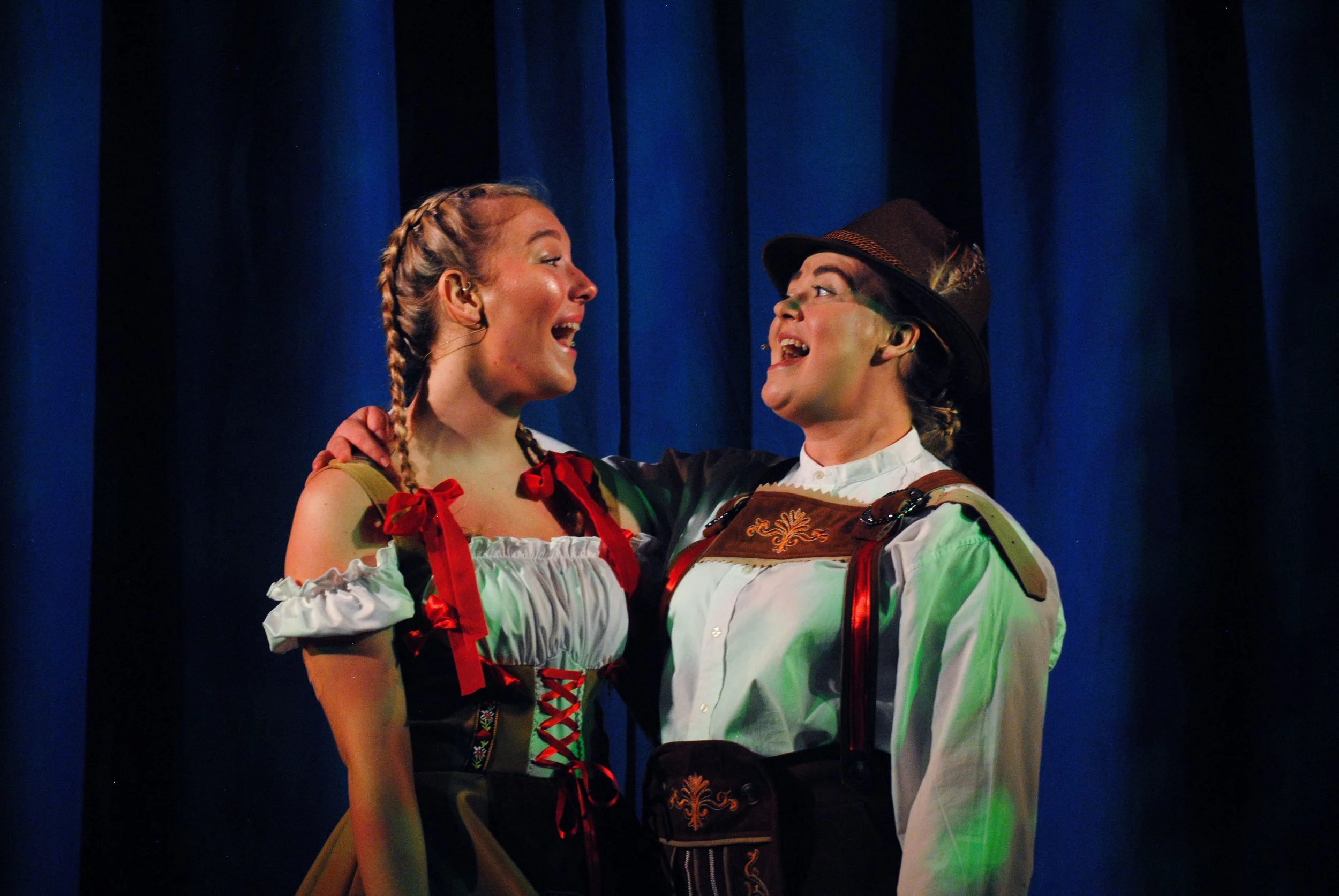 Two women in traditional Bavarian costumes singing on stage against a dark blue curtain.