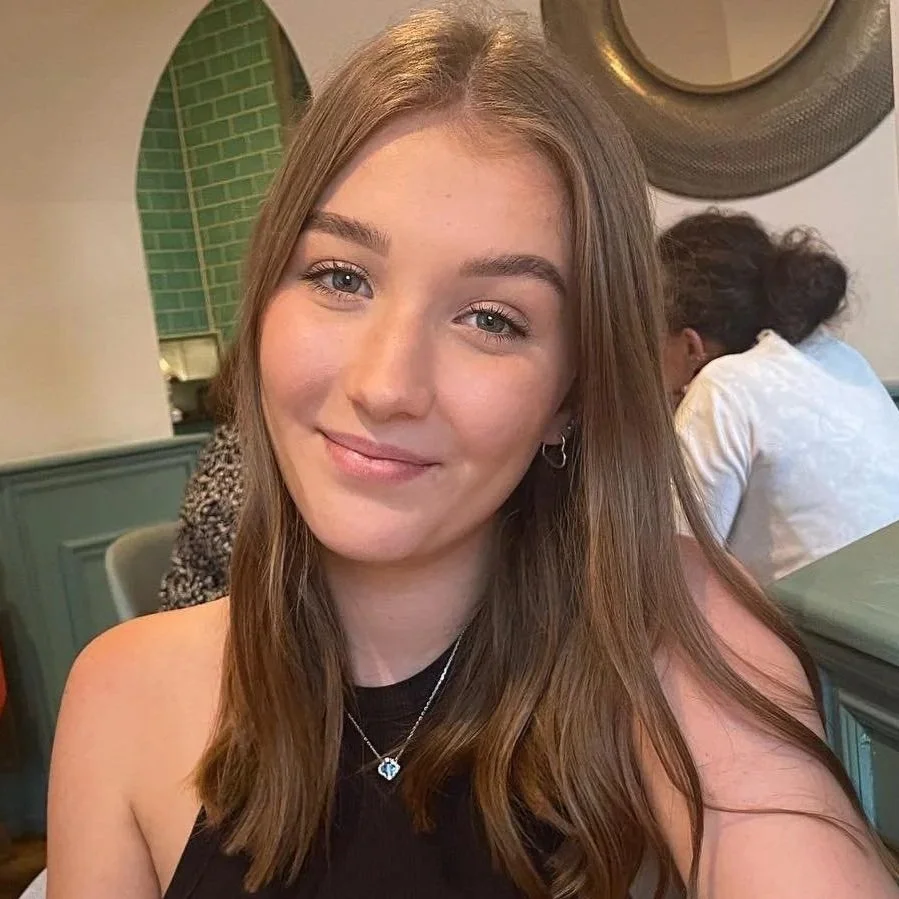 A smiling young woman with shoulder-length brown hair, wearing a black top, earrings, and a necklace with a blue pendant, sitting in a restaurant.