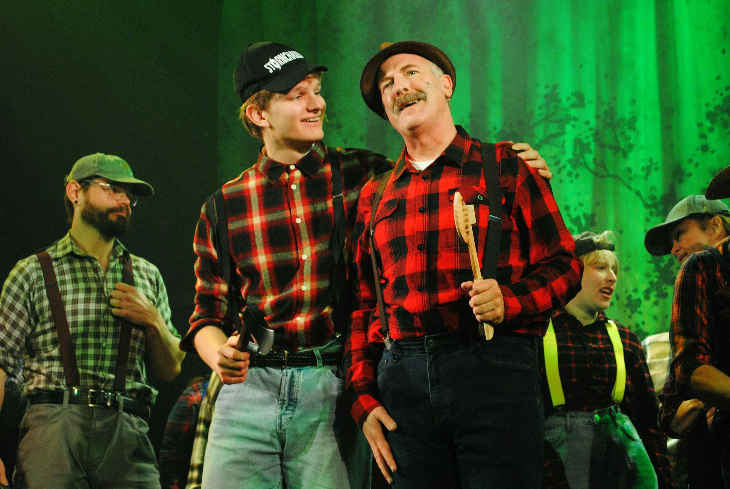 Actors performing on stage in a theatrical play with a forest background, dressed in plaid shirts and hats, including a man holding a wooden spoon, emphasizing a rustic or rural scene.
