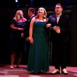 Four people, two women and two men, standing together on a stage, engaging with each other; formal attire suggests a performance or event.