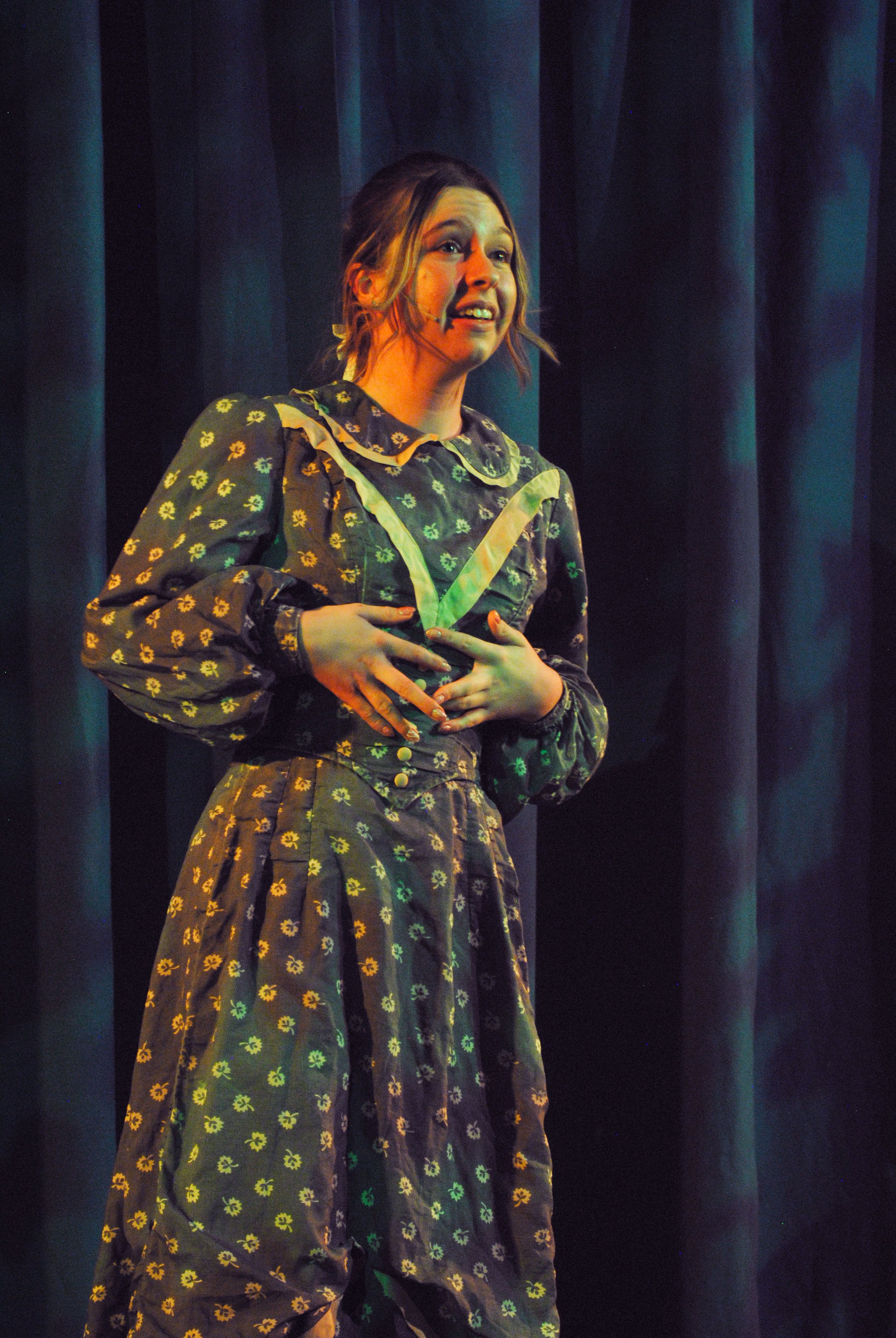 A woman on stage dressed in a vintage-style dress with a floral pattern, holding her stomach and speaking or singing, with dark blue curtains in the background.
