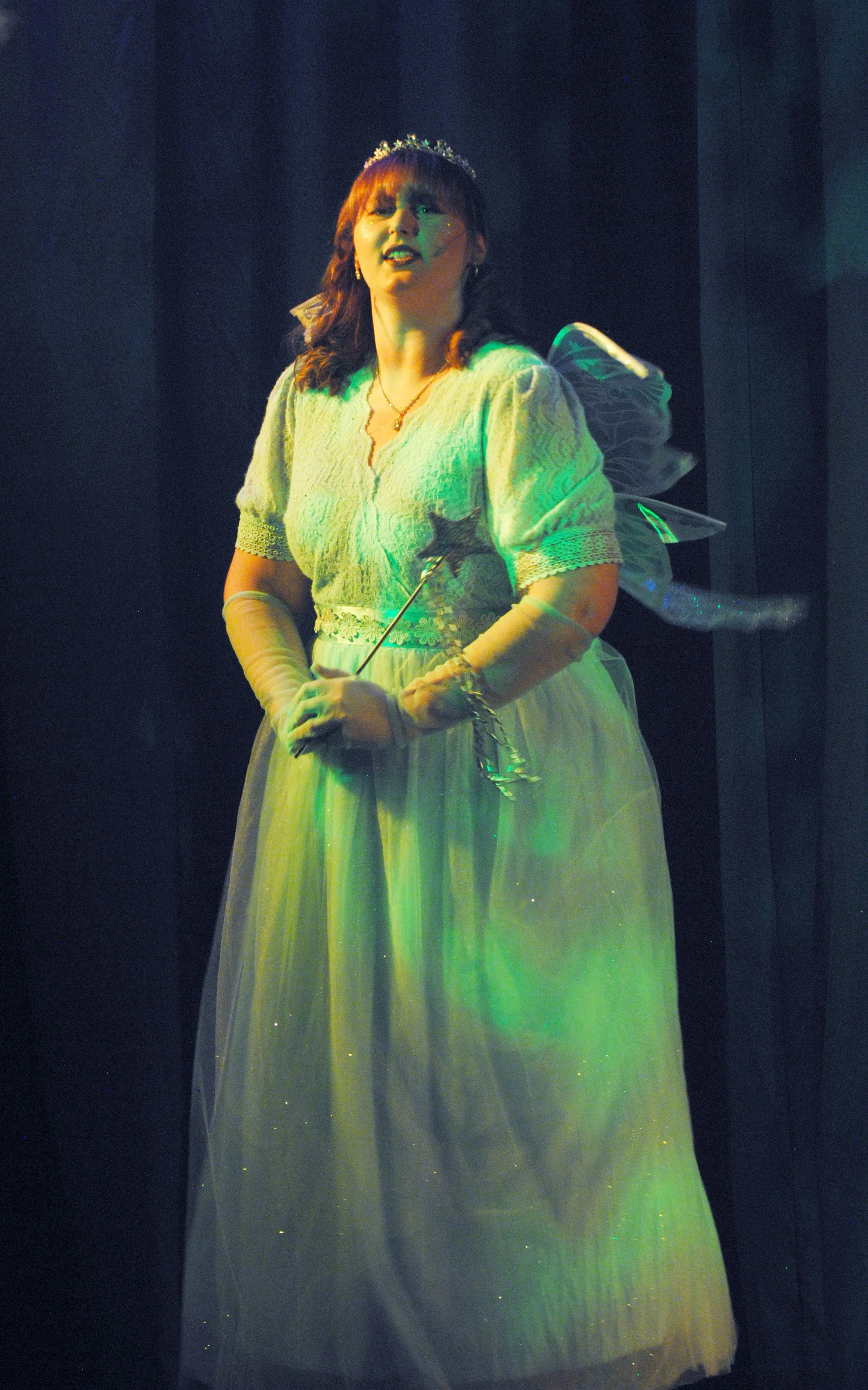 A woman dressed as a fairy princess, holding a wand, wearing a crown and fairy wings, standing on a stage with dark curtains.
