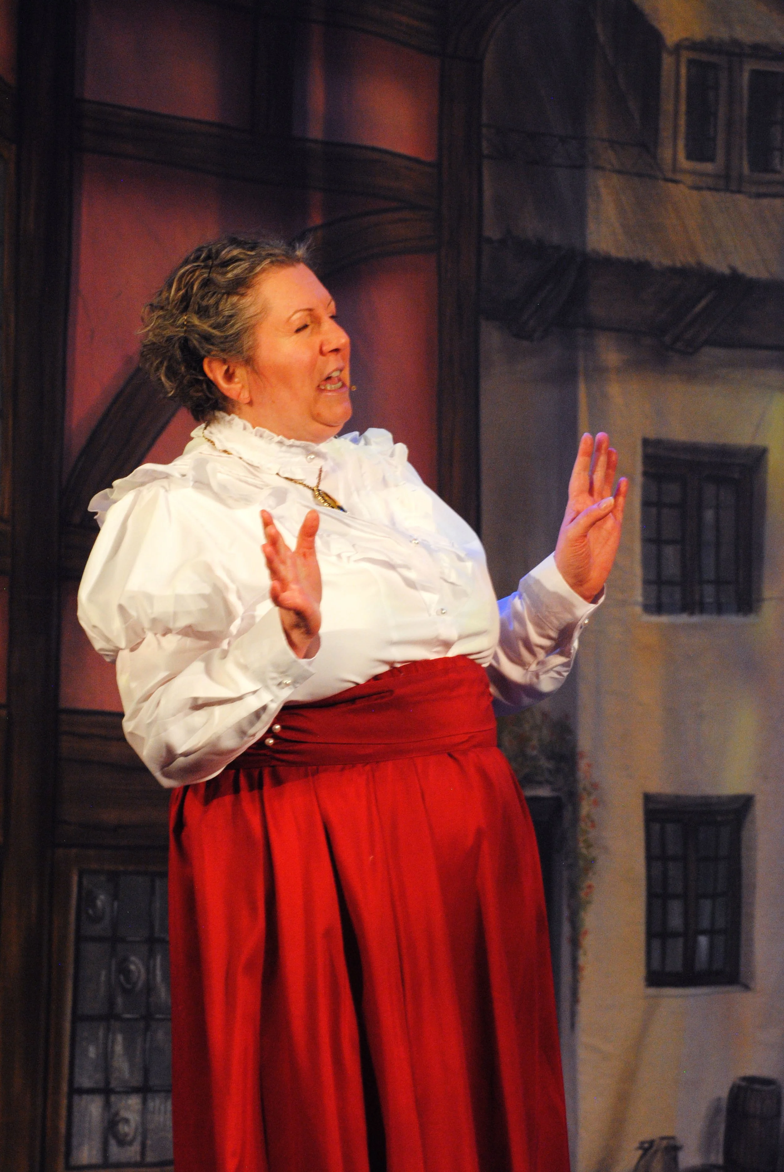 A woman dressed in a white blouse with ruffled collar and red skirt, performing on stage with a painted backdrop of old buildings.