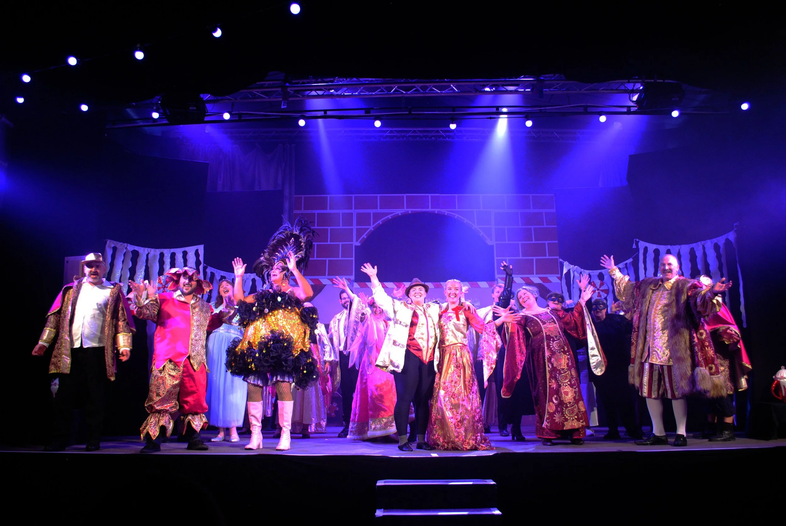 A stage performance featuring actors in colorful costumes, some with feathered hats and ornate robes, bowing with their arms raised, with a painted backdrop resembling a castle wall and lanterns hanging above, under blue lighting.