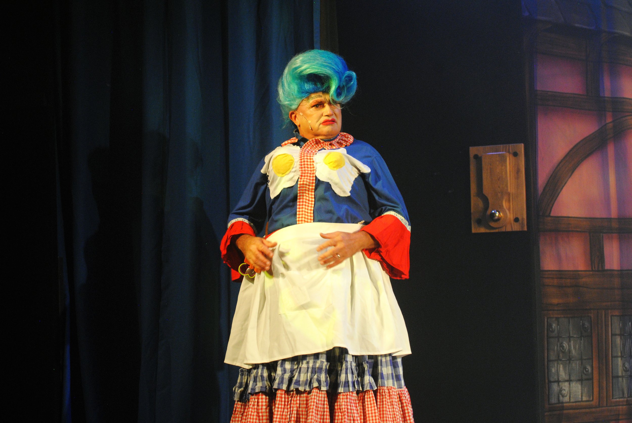 A performer dressed as a clown with a blue and green wig, red lipstick, and a colorful costume with floral and checkered patterns, standing on stage with a dark curtain background.