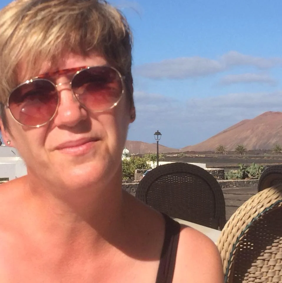 A woman with short light brown hair, wearing sunglasses, smiling outdoors with a mountain and a blue sky with some clouds in the background.
