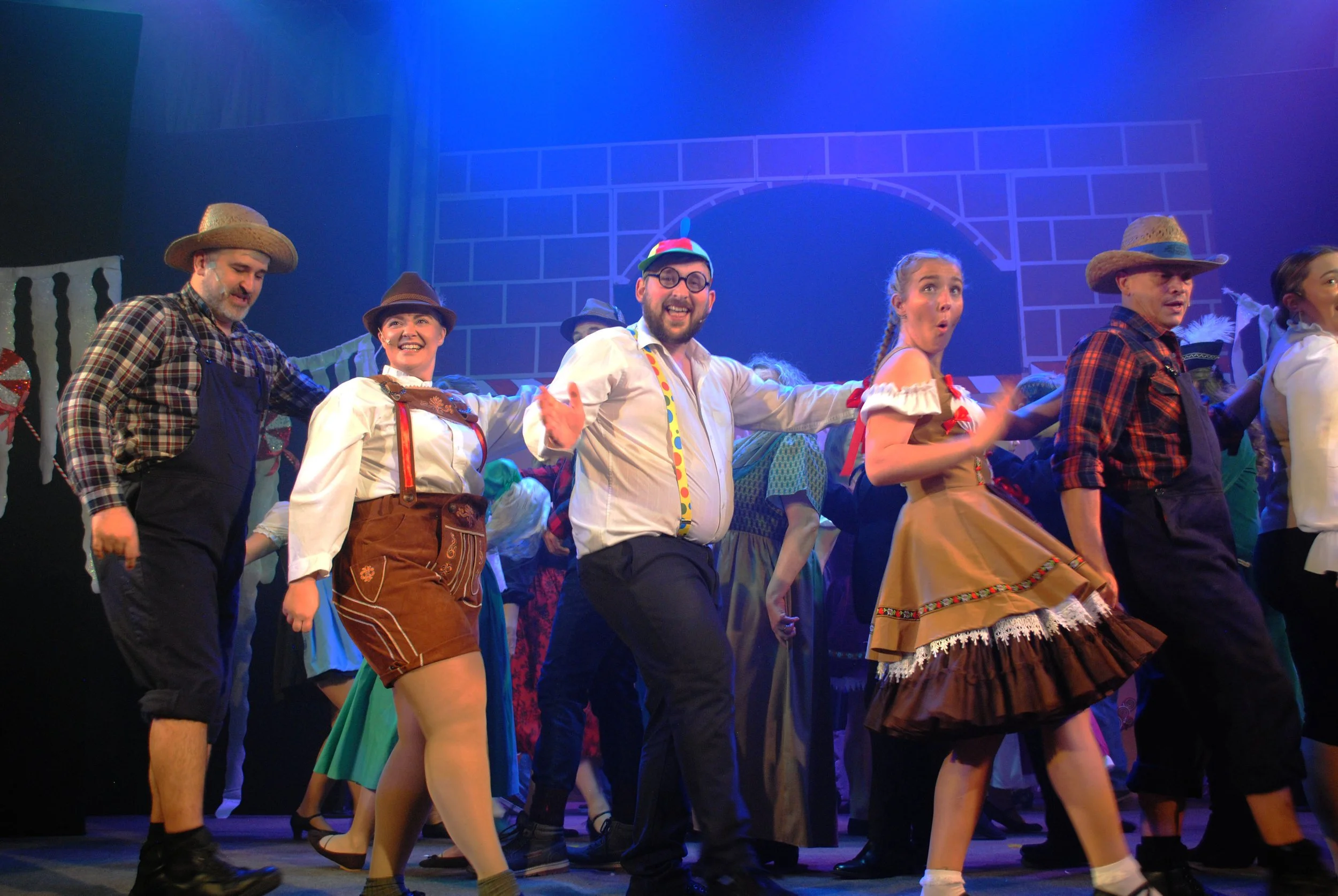 A lively stage performance with actors in colorful costumes, some in traditional European folk attire and others in casual clothing, smiling and dancing under stage lighting with a castle-like backdrop.